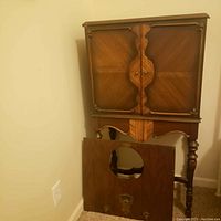 Front view of antique wooden cabinet with distinctive paneling and carved legs, showing the front door panel removed revealing the empty interior where the radio once was.