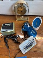 Overview of all four clocks on wooden floor including Seiko mantel clock, digital alarm clocks, and blue drawstring pouch.