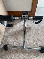 Pedal exerciser with metal frame, black foot pedals, and straps shown on carpet.