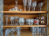 Full view of wooden shelves holding various glassware items including a decanter, multiple types of wine glasses, tumblers, and decorative glasses with floral accents. Items appear unused for some years, with some needing washing.