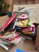 Pile of assorted kitchen linens including floral pot holders, cloth napkins in plastic packaging and loose, and folded fabric pieces possibly tablecloths.