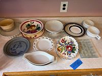 Photo showing an assortment of 12 assorted dishes and bowls on wooden counter, with various patterns and colors visible, including fruit, floral, and solid colors.