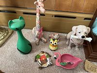 Overview of six animal figurines and decor items on carpeted floor including cat, giraffe, owl, dog, turtle, and flamingo shaped dish.