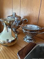 Photo showing a reflective silver-tone vase, ornate sugar bowl with lid, and matching creamer with detailed engraving on wooden surface.