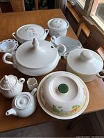 View of entire lot showing various covered bowls, tureens, teapot, and other ceramic kitchenware arranged on wooden table.