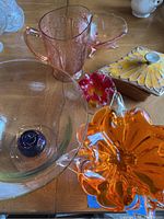 Photo showing a collection of various artistic glassware pieces and a ceramic bowl with a floral lid on a wooden surface.