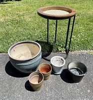 Photo of the collapsible metal plant stand with wooden tray top alongside one large glazed ceramic pot and four smaller ceramic and terracotta pots arranged on asphalt with grass background.