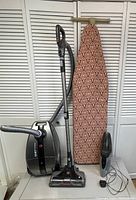Photo showing Hoover canister vacuum with wand and hose, Shark cordless handheld vacuum with charging plug, and upright ironing board with patterned cover.