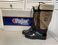 Pair of women's Pajar Canada winter boots showing glossy black waterproof lower, beige suede upper with wool cuff, positioned in front of original brand box