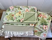 Image shows a folded full/queen reversible floral quilt with green striped reverse, two matching standard quilted floral shams, and a light gray polyester blanket with satin edges arranged on a table.