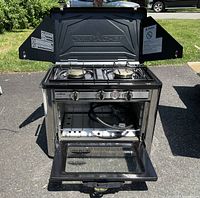Open view of the Webber Camp Chef propane oven showing two burners on top, control knobs, oven cavity with hose inside, and oven door open.