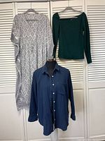 Photo of three clothing items hanging on a wall: grey and cream caftan on left, green long sleeve asymmetrical neckline t-shirt on right, navy button down shirt below, displayed on mannequin torso.