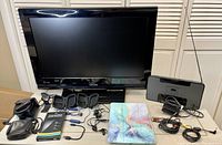 Photo shows a Toshiba flat screen TV approx 26" with power cord, black Altec Lansing speaker/radio with AUX cord and stand for older iPhones, multiple headphones and cables laid out, iPad cover with colorful front, various cases including camera cases and boxes for iPhone screen protectors.
