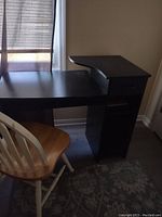 Black small computer desk placed near window with light filtering through blinds, showing main desk surface, raised side shelf, drawer and single cabinet door beneath.