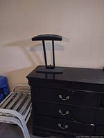 Black desk lamp placed on a black dresser against a white wall, showing the full lamp from base to shade.