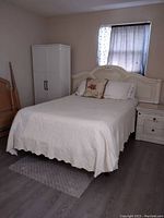 Queen-sized bed featuring a cream-colored headboard with carved panel design. Adjacent to the bed is a matching cream lamp stand/nightstand with two drawers and a cabinet. The room has a light-colored floor and a window with curtains.