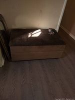 Photo of brown fabric ottoman with textured top and storage capacity, placed against a wall in a room.
