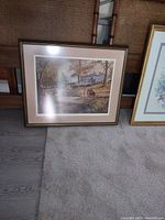 Framed landscape picture showing an autumn scene with trees, water, and buildings, resting on the floor against a wall.