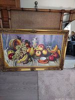 Front view of the still life fruit print picture in an ornate gold frame leaning against a chair with cane back and amidst other furniture items.