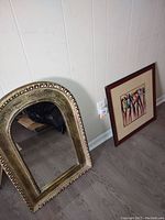 Photo showing the framed artwork of black dancers leaning against the wall next to the ornate gold framed mirror.