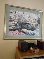 Framed landscape painting depicting flowers in bloom, a pond, and leafless trees, hung on a wall above wooden speakers.