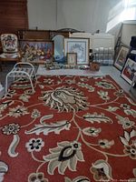 Photo of a large floral area rug with a vibrant red-orange background and beige, cream, and brown flowers and leaves, situated on a floor with various frames and furniture in the background.