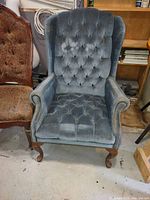 Front view of wing chair with blue velvet tufted fabric, wooden cabriole legs and nailhead trim on armrests. Chair is set on a concrete floor near another upholstered chair and wooden furniture.