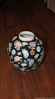 Front angled view of a large Chinese jar with colorful floral patterns on a black base placed on a wooden floor.