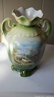 Full view of green porcelain vase showing two handles, scalloped white top rim, and rural landscape painting on the body.