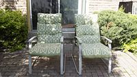 Front view of the pair of outdoor patio chairs with green cushions placed on a brick patio in front of a door and surrounded by green shrubbery