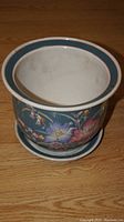 Photo showing top-down view of ceramic plant pot with white interior, blue and white rim banding, and floral exterior design, set on wooden floor.