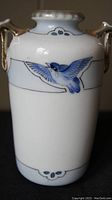 Front side of a white porcelain vase decorated with a flying blue bird and gold-tone handles.