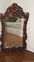 Full view of large wood framed mirror standing on wooden floor, showing entire frame and some background furniture.