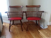 Two red Windsor chairs positioned side by side on a hardwood floor, showing full front views highlighting wooden spindle backs and red leather seats.