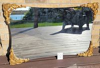 Mirror displayed horizontally with decorative golden corner frames and reflective glass surface showing outdoor background.