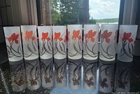 Ten frosted glasses with orange flower designs lined up in a row on a reflective surface with natural light showing details of the design.