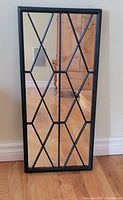 Front view of the rectangular mirror with black metal geometric overlay pattern, placed against a wall on a wooden floor.