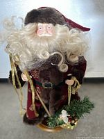 Front view of porcelain faced Santa figure showing detailed face, curly white beard and hair, red and brown velvet attire with fur trim, holding a staff and greenery bundle.