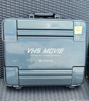 Closed carrying case showing 'VHS Movie Integrated Camera/Recorder Hitachi' branding on top surface. The case is hard plastic with handle.
