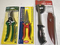 Photo shows all three items new in package: pruner with green handles, scissors with red and yellow handles, and Eagle Claw fillet knife with leather sheath.