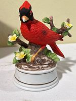 Close-up photo of one red cardinal bird music figurine, perched on a branch with yellow flowers and green leaves, on a white base.