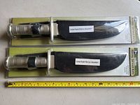 Pair of packaged hunting/survival knives with black sheaths and plastic handles on top of the packaging with measuring tape showing about 8-inch blade length