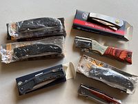 Seven folding pocket knives in original boxes or plastic bags arranged on table.