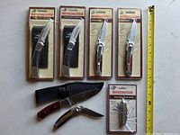 Seven Winchester folding knives displayed, five sealed in original packaging, two unused outside packaging. Two black rubber inlay knives with sheaths, two wood inlay serrated blade knives with pocket clips sealed, one sealed Swiss style multi-tool knife, and two unused wood inlay folding knives with sheaths outside packaging.