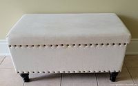 Front view of light beige fabric storage bench with studded trim and black legs on tiled floor.