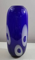 Front view of a tall cylindrical cobalt blue glass vase with white oval 'eye' decorations spaced evenly around the body.