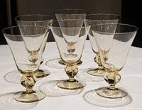 Six Victorian flint water goblets arranged in two rows on a white surface, showing the clear glass bowls and pale gold blown stems with footed bases.