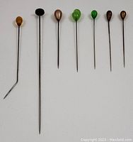 Full view of 7 vintage hat pins in a row showing various colors and shapes of decorative tops on slender metal shafts.