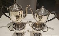 Photo showing tea pot, coffee pot, open sugar bowl, and creamer in polished EP copper with lead mounts, arranged on a reflective surface.