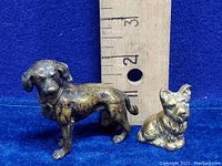 Two vintage brass dog figurines placed next to a ruler for size reference, showing approximate heights of about 2.5 inches and smaller.
